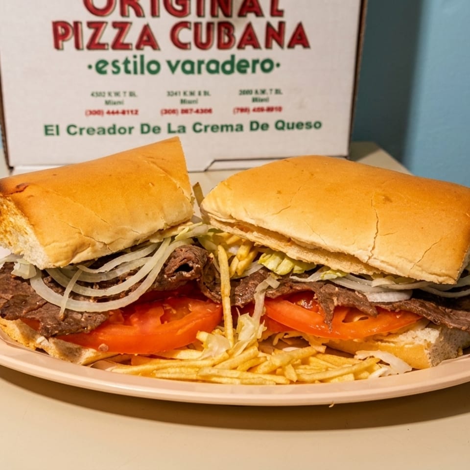 A Cuban steak sandwich with onions, tomatoes, and shoestring fries on a plate, with a pizza box in the background.