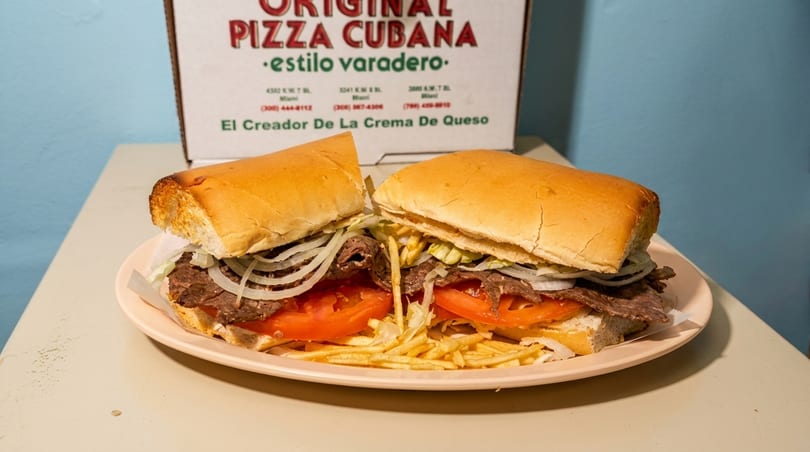 A Cuban steak sandwich with onions, tomatoes, and shoestring fries on a plate, with a pizza box in the background.