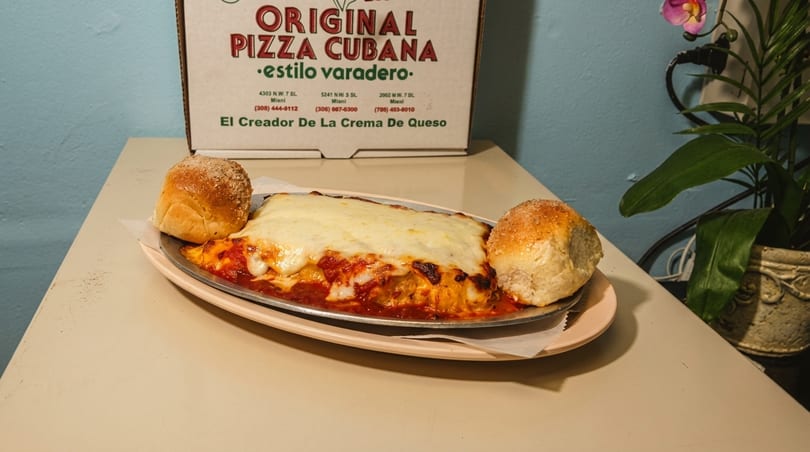 Baked lasagna with melted cheese and two bread rolls, next to an Original Pizza Cubana box.