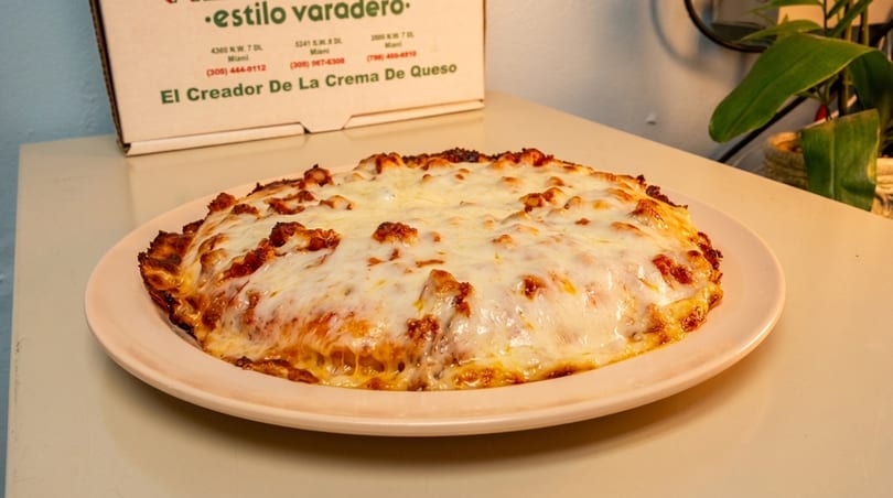 A cheesy, baked dish, possibly lasagna or a casserole, on a light pink plate with a pizza box in the background.